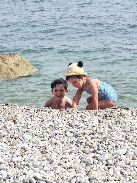 A little boy and girl smiling happily and playing in the waves on a white stone beach on Italy's Amalfi Coast.
