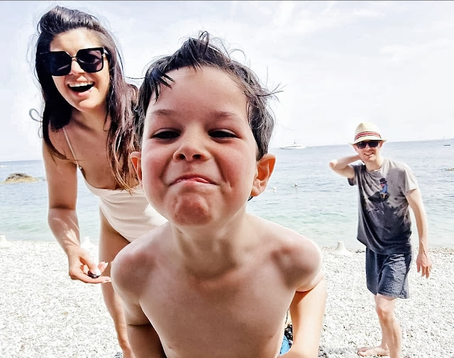 A happy family smiling on a white stone beach in Italy - a mother in a white swimsuit and sunglasses, a dark haired little boy pulling a cheeky face at the camera, and a father in damp swim shorts with a straw fedora hat.
