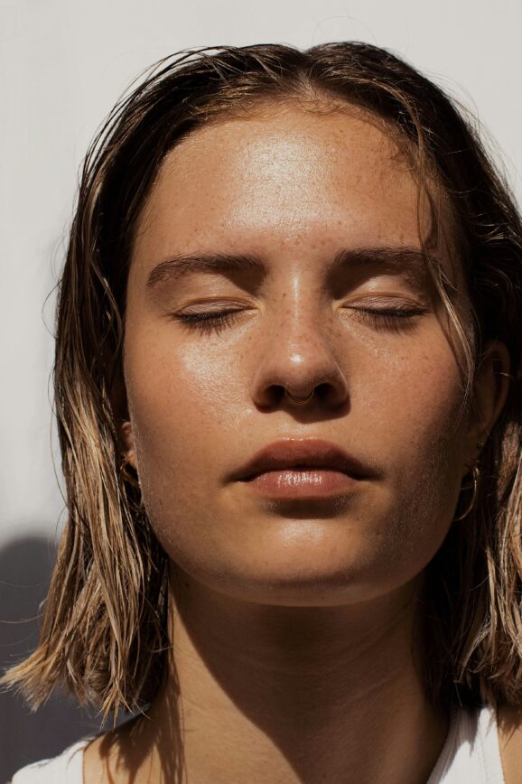Woman with dewy, glowing skin and closed eyes basking in sunlight; minimalist summer beauty look with natural makeup and wet-look hair.