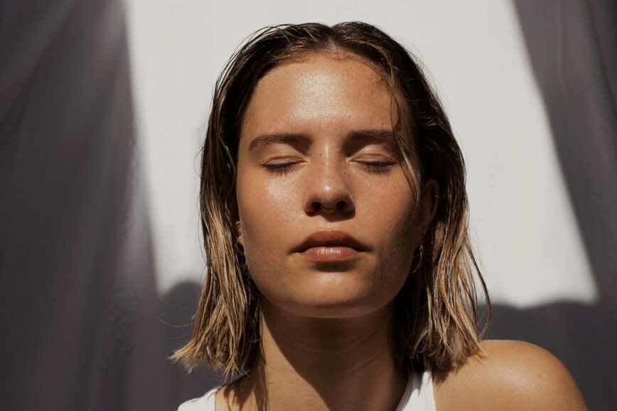 Woman with dewy, glowing skin and closed eyes basking in sunlight; minimalist summer beauty look with natural makeup and wet-look hair.